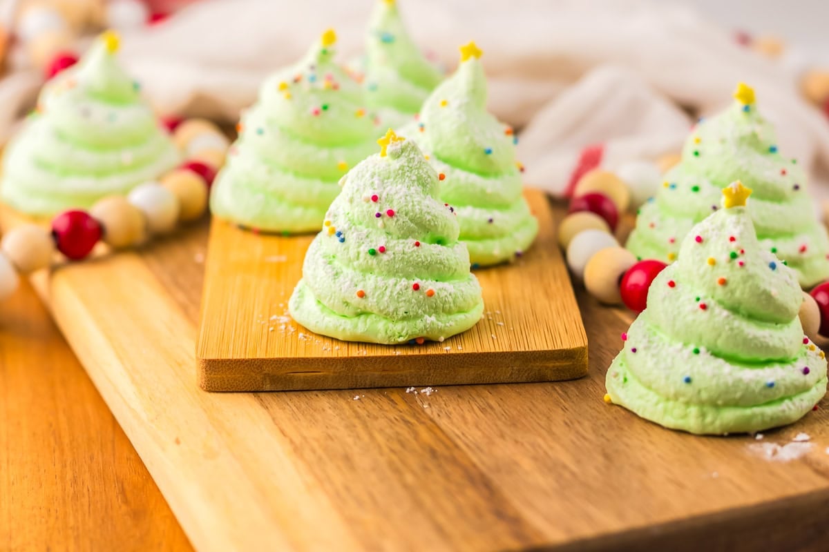 Green meringue cookies shaped like Christmas trees with colorful sprinkles and yellow star decorations, arranged on a wooden board.