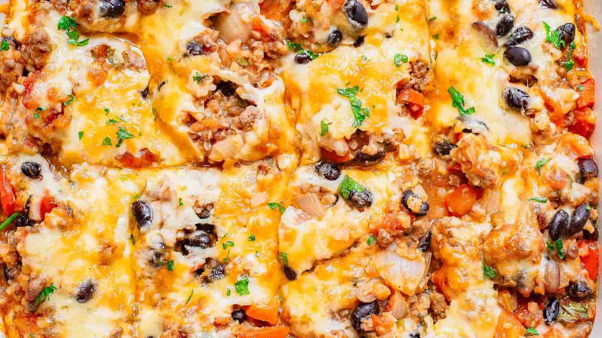 Close-up of a baked casserole topped with melted cheese, ground meat, black beans, onions, and chopped parsley, cut into square portions.