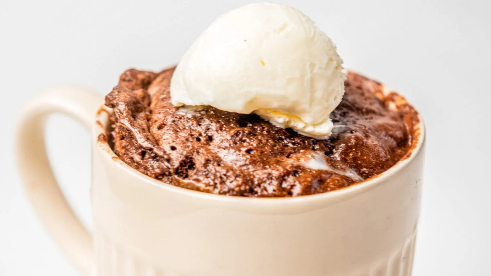 A mug filled with chocolate mug cake topped with a scoop of vanilla ice cream, set against a plain background.