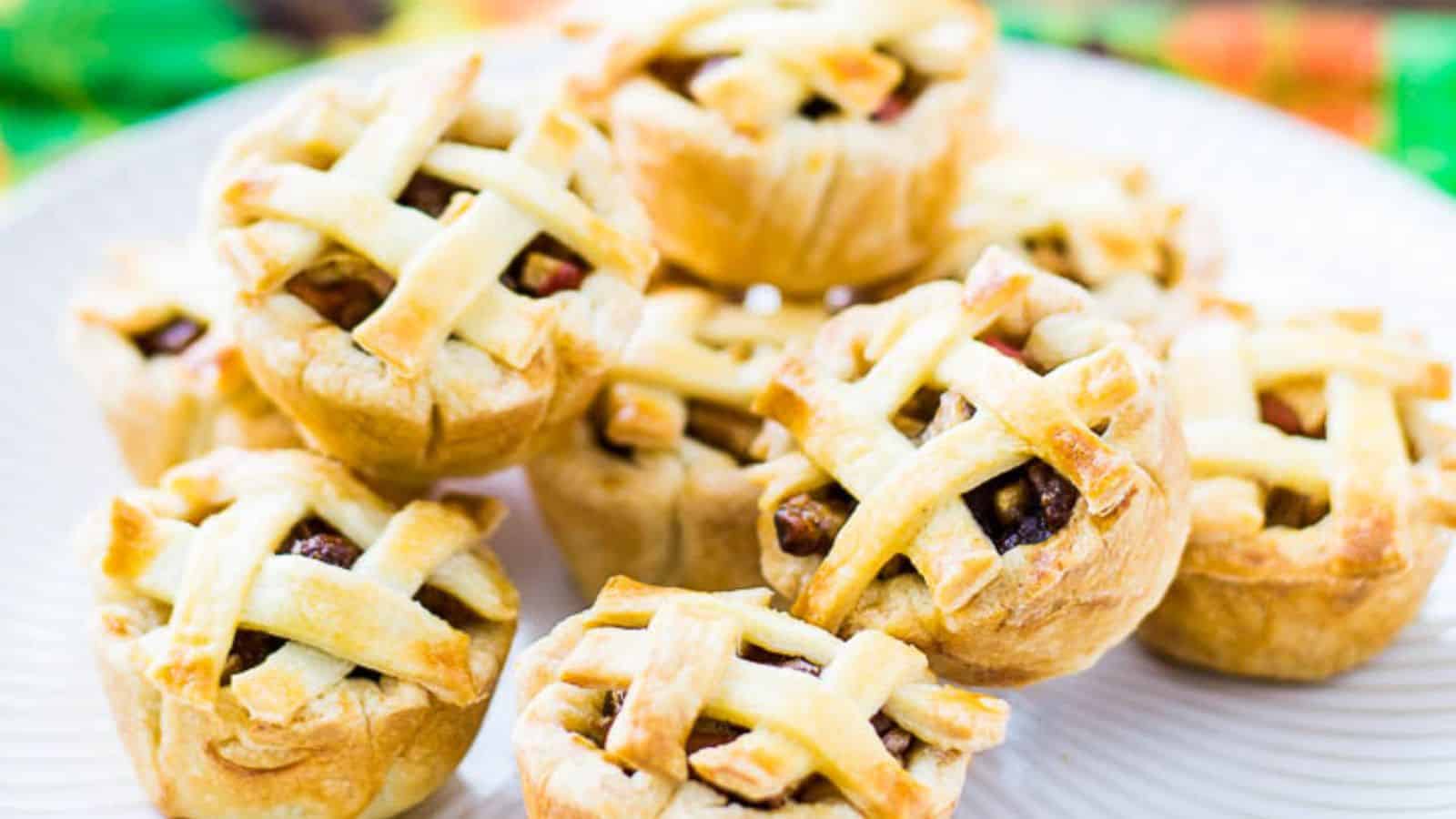 A plate of mini lattice-topped apple pies arranged in a pile, with flaky golden crusts and visible apple filling.