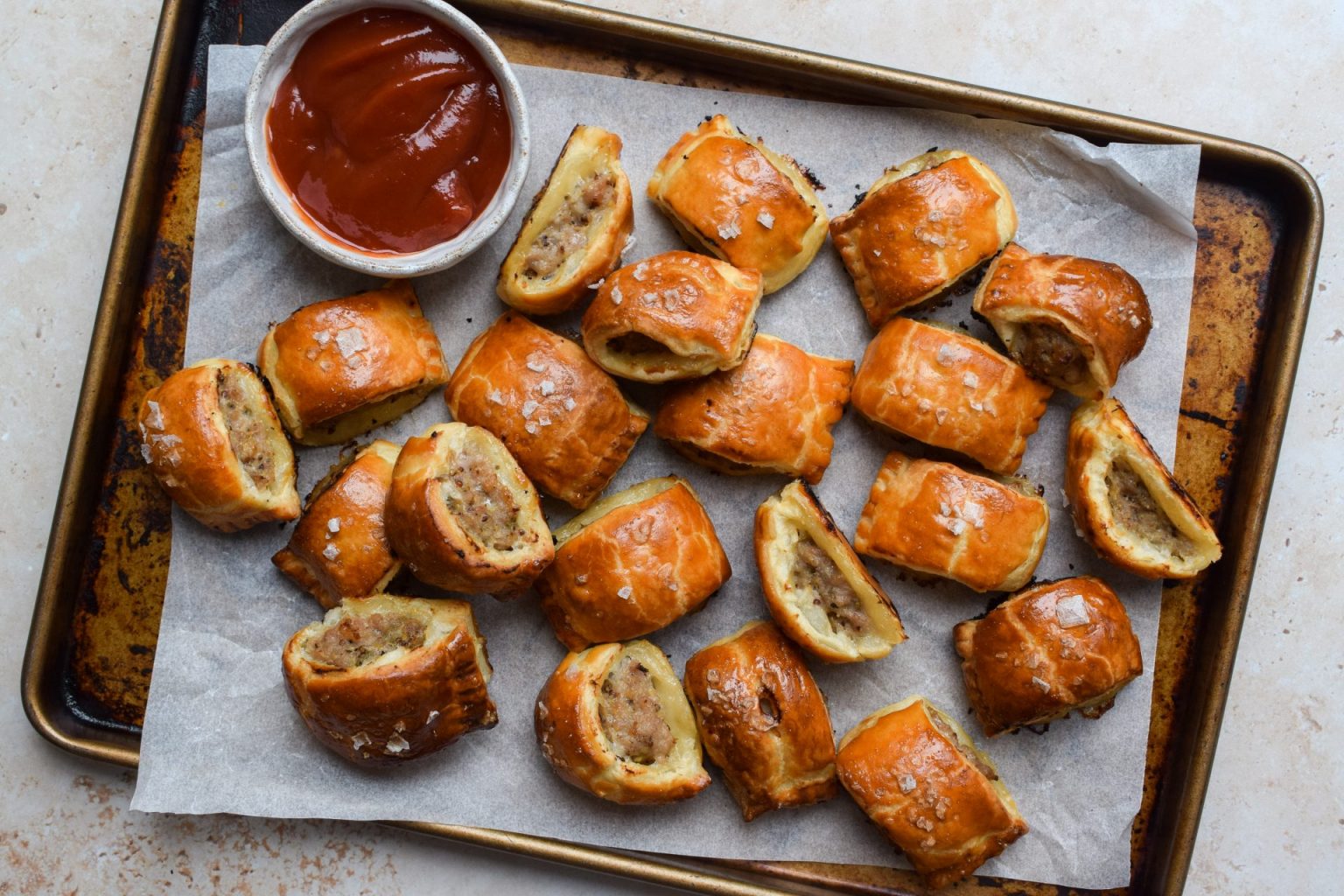 A baking tray with several sausage rolls on parchment paper, served with a small bowl of red dipping sauce.
