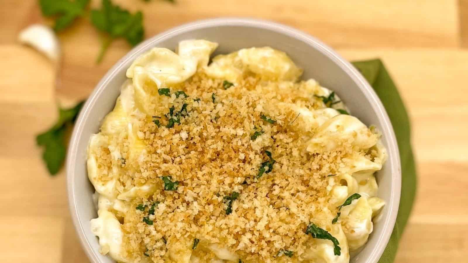 A bowl of macaroni and cheese topped with golden toasted breadcrumbs and chopped herbs, placed on a wooden surface.