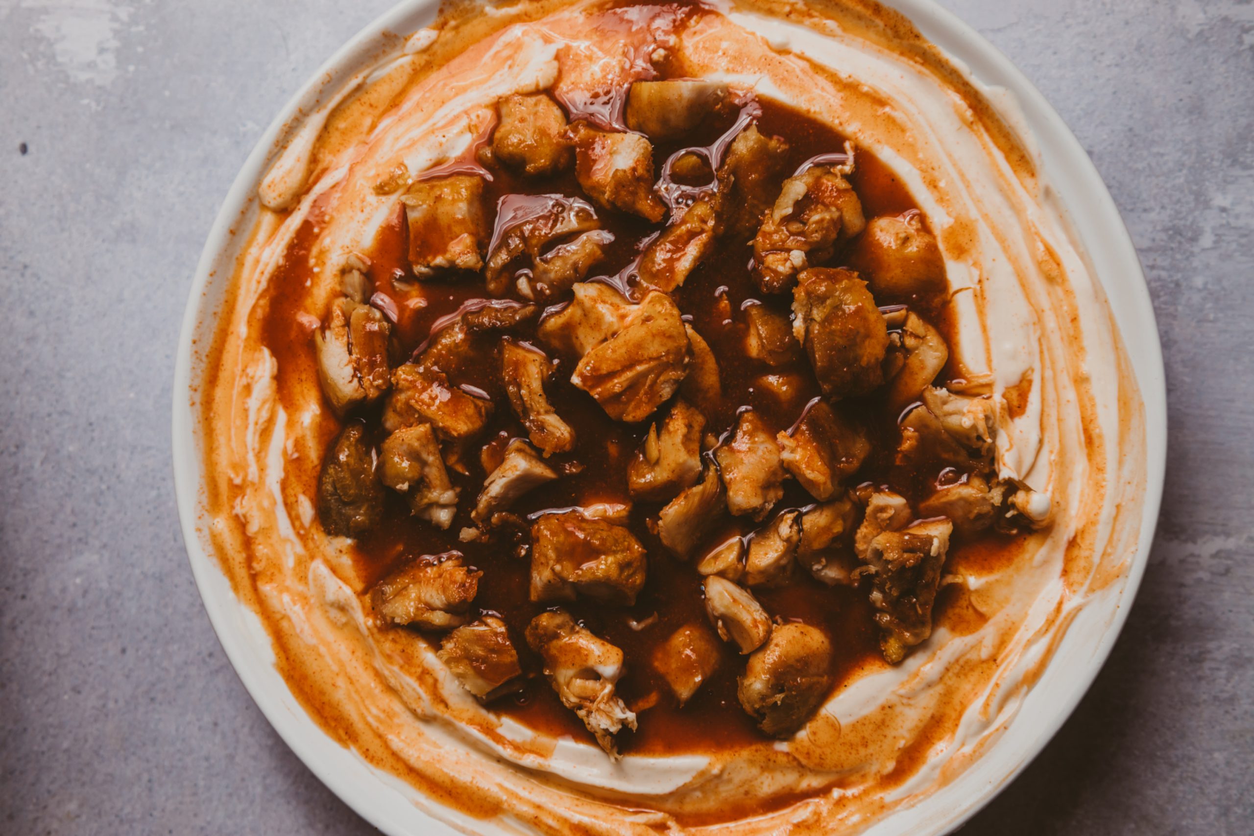 A white plate with chunks of meat in a red sauce, served on a base of creamy white spread, on a light gray surface.