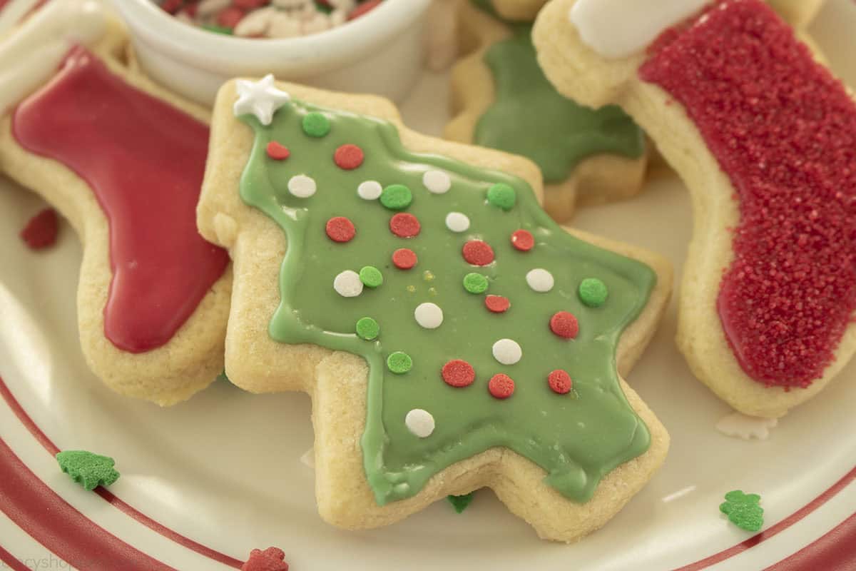 A plate of decorated Christmas cookies, including a green Christmas tree with red, white, and green sprinkles and a red stocking-shaped cookie.