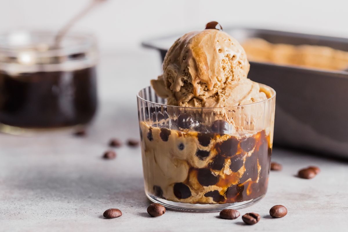 A clear glass cup filled with coffee ice cream, tapioca pearls, and dark syrup, with coffee beans scattered on the surface nearby.