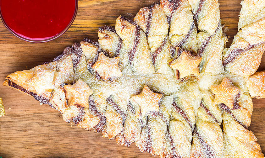 A Christmas tree-shaped pastry decorated with star-shaped pieces and powdered sugar, served on a wooden board with a glass of red beverage nearby.