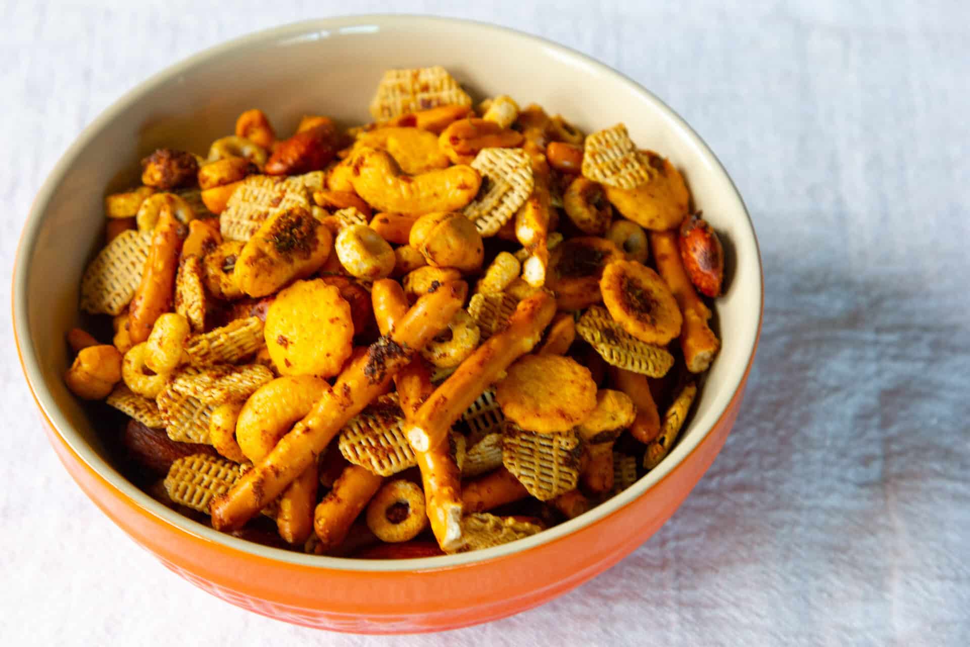 A bowl of snack mix containing pretzel sticks, cereal squares, cheese crackers, nuts, and other bite-sized pieces on a white surface.