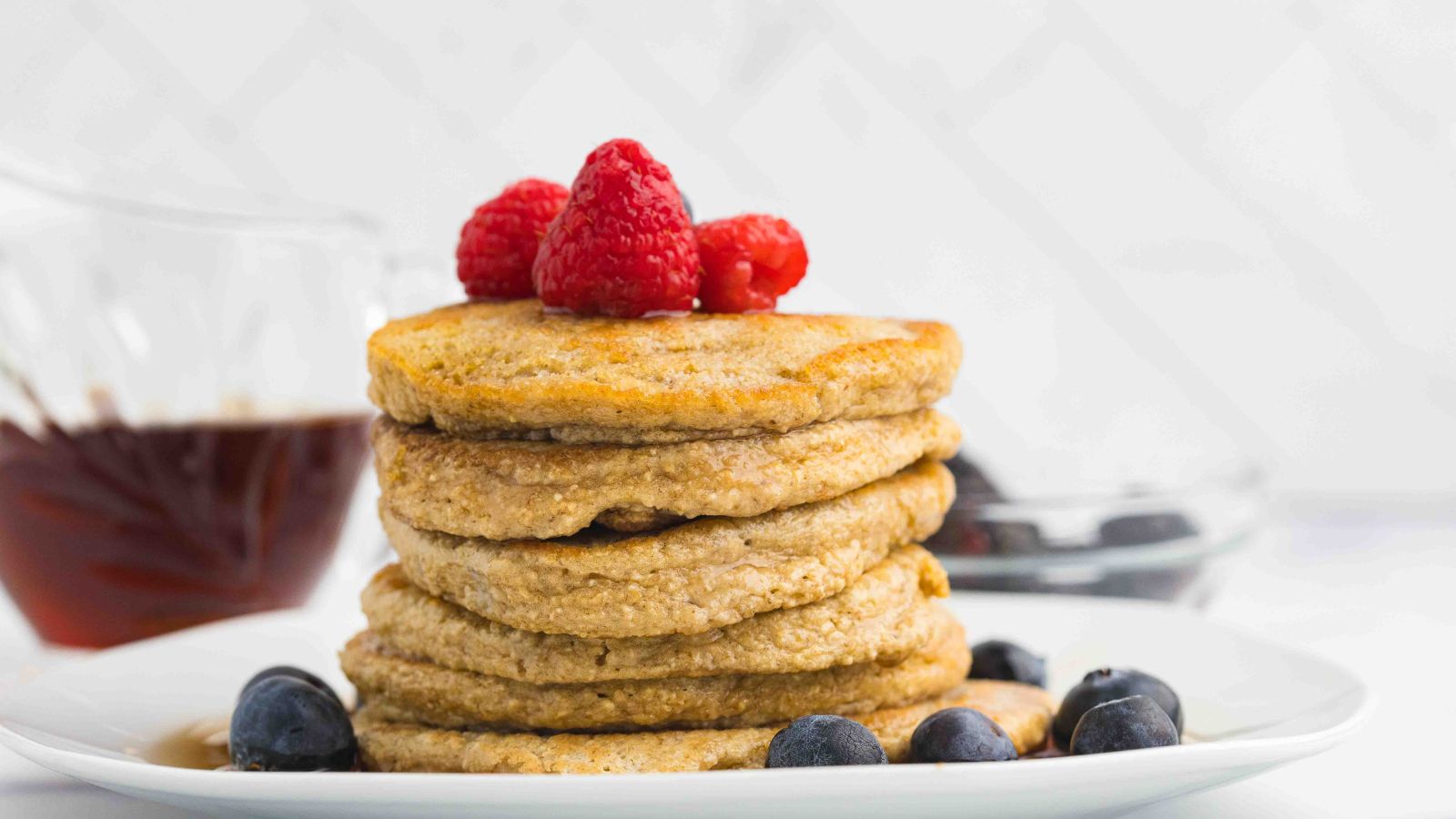 A stack of pancakes topped with raspberries and surrounded by blueberries on a white plate, with a glass container of syrup in the background.