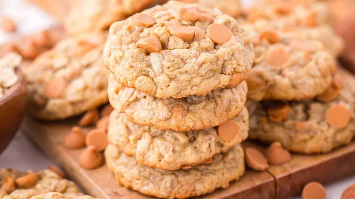 A stack of oatmeal cookies with butterscotch chips on a wooden board, with more cookies and chips scattered around.