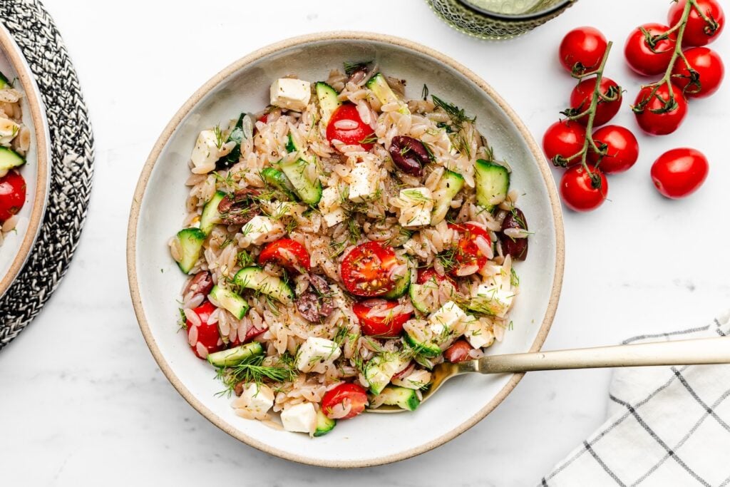 A bowl of orzo salad with cherry tomatoes, cucumber, feta cheese, olives, fresh dill, and a fork, placed on a marble surface next to a bunch of vine tomatoes.