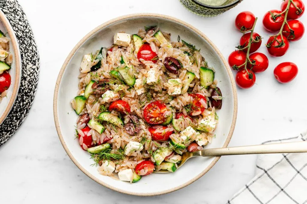 A bowl of orzo salad with cherry tomatoes, cucumber, feta cheese, olives, and dill, served with a gold-colored fork.
