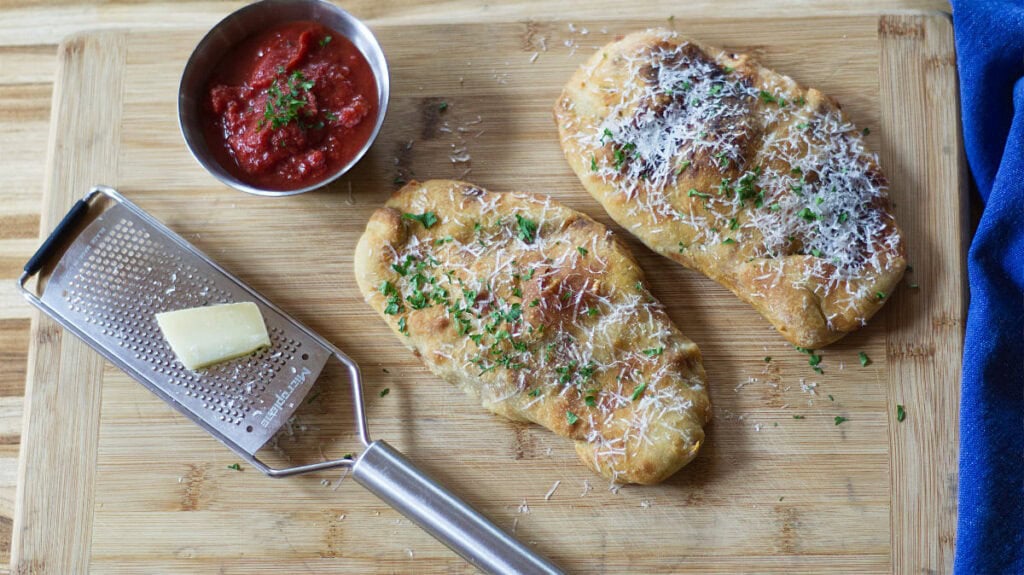 Two pieces of cheesy bread topped with herbs and grated cheese are on a wooden board, next to a metal grater, a block of cheese, and a bowl of red dipping sauce.