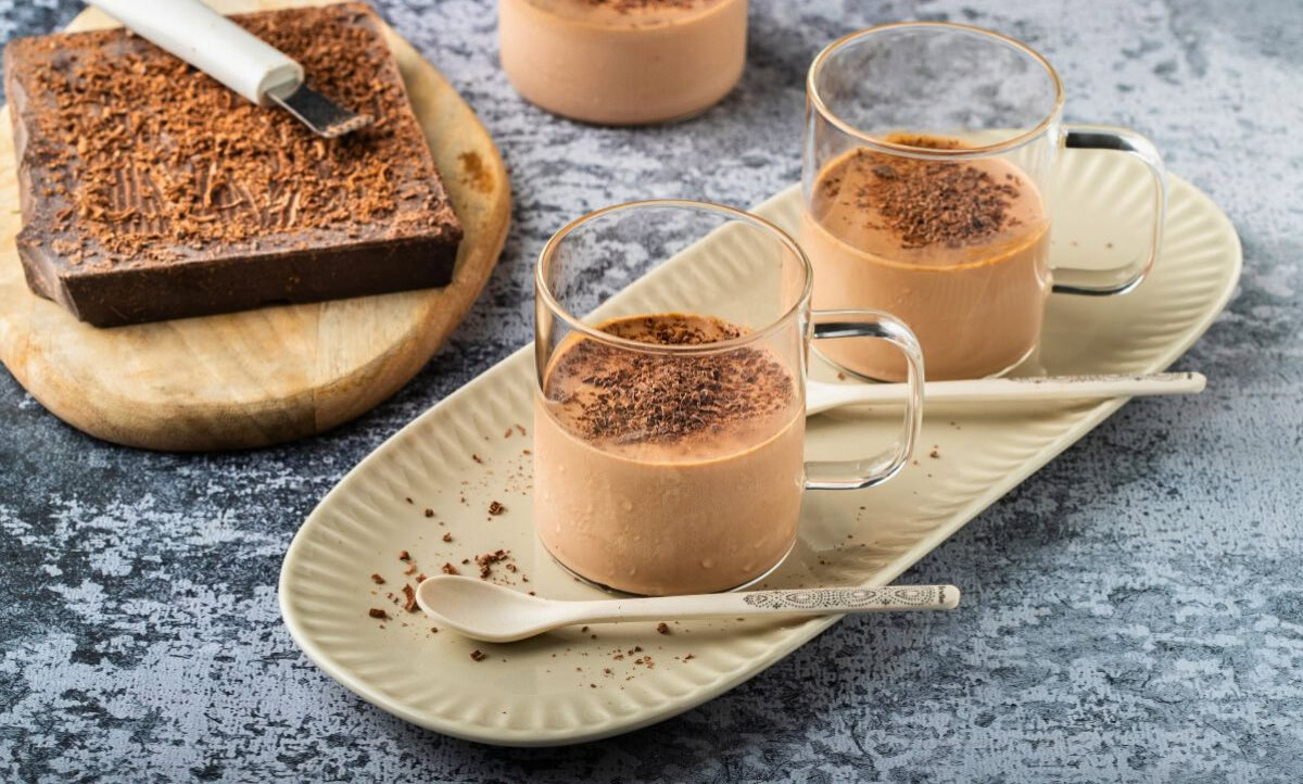 Two glass cups of chocolate mousse topped with chocolate shavings on a beige plate with spoons, next to a block of chocolate on a wooden board.