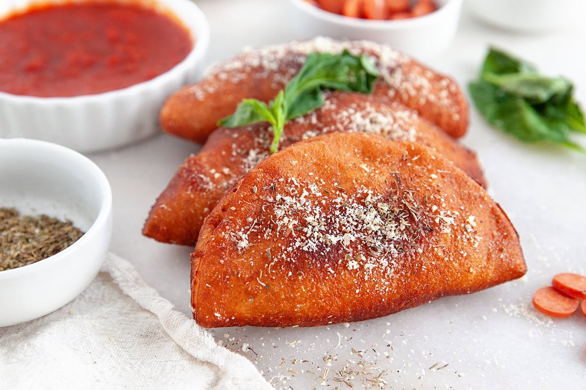 Three fried calzones topped with grated cheese and herbs, garnished with fresh basil, are arranged on a white surface next to bowls of marinara sauce and dried herbs.