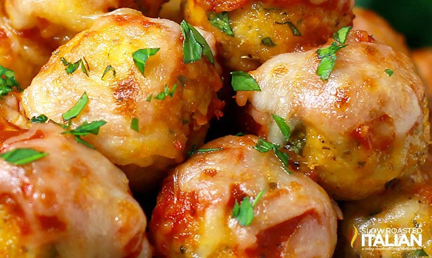 Close-up of baked meatballs topped with melted cheese, tomato sauce, and sprinkled with chopped parsley.
