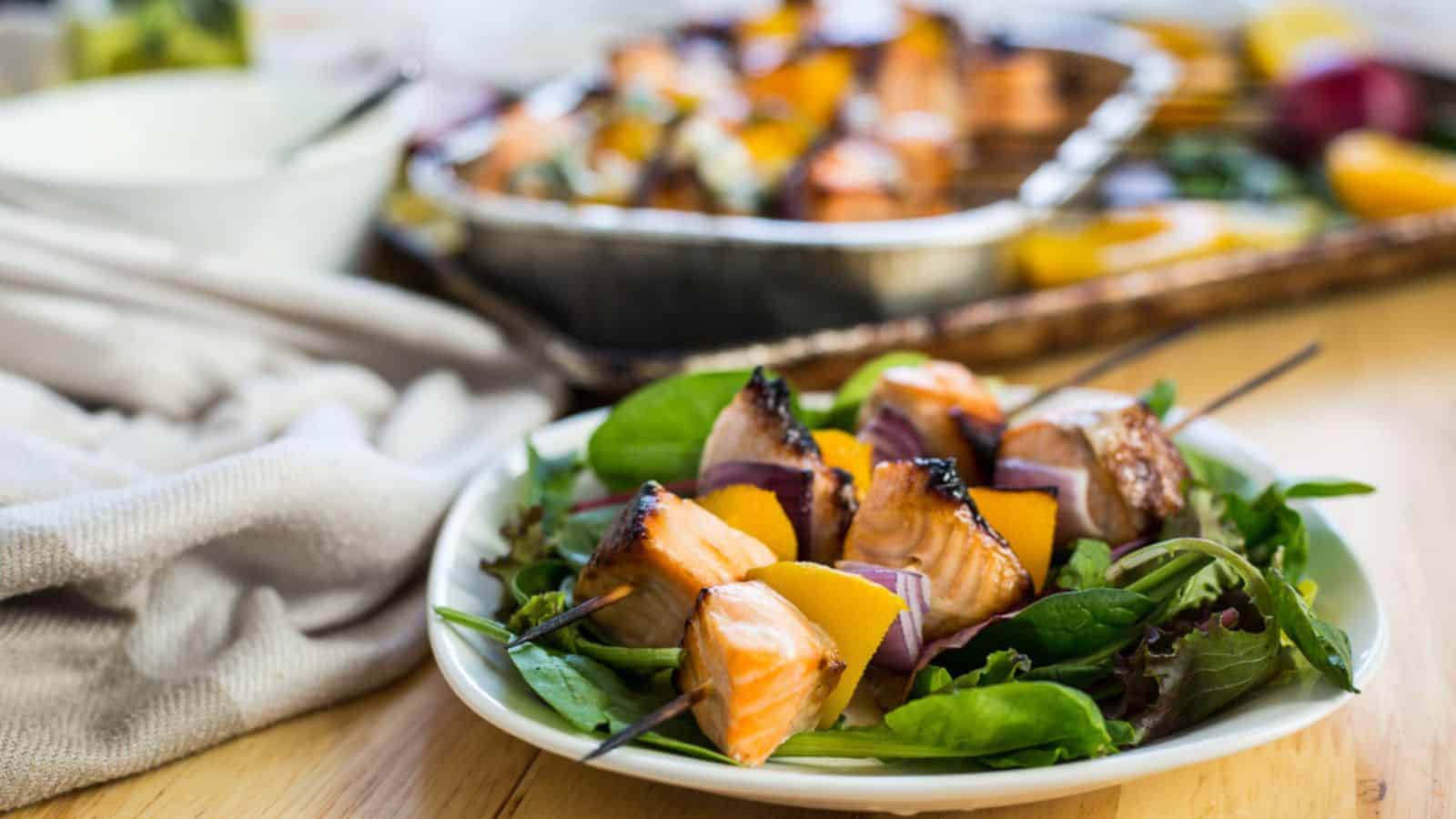 A plate of grilled salmon, red onion, and yellow pepper skewers served on a bed of mixed greens, with a tray of more skewers in the background.