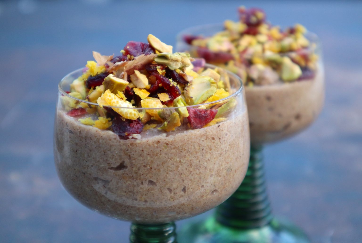 Two glass dessert cups filled with a creamy brown pudding, topped with chopped pistachios and dried fruit, are displayed against a blurred background.