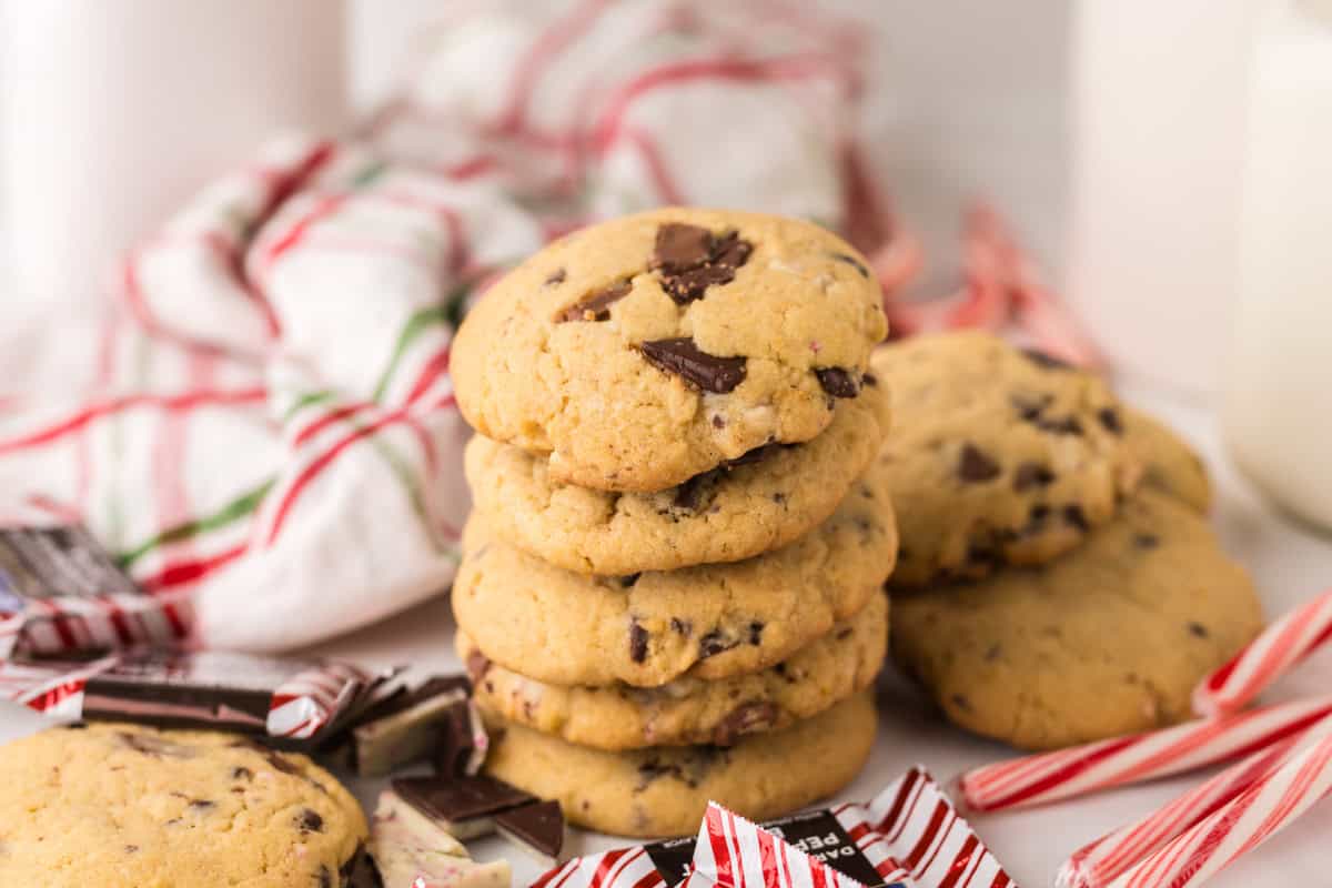 A stack of chocolate chip cookies sits on a white surface, surrounded by peppermint candies, with a red and white striped cloth in the background.