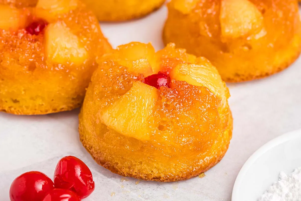 Mini pineapple upside-down cakes topped with pineapple chunks and a cherry, with additional cherries on the side, displayed on a light surface.