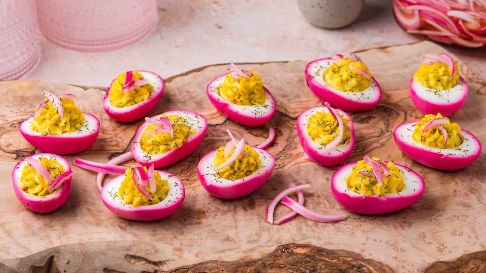 Deviled eggs with bright pink outer whites, topped with yellow filling and garnished with pickled onions and herbs, arranged on a wooden serving board.