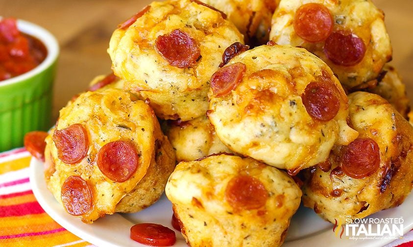 A plate of pizza muffins topped with mini pepperoni slices sits on a table, with a small green bowl of marinara sauce in the background.