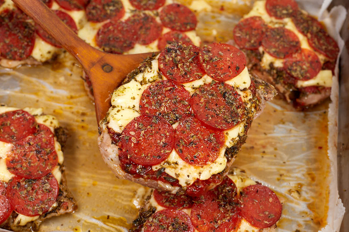 A wooden spatula lifts a slice of baked chicken topped with melted cheese, pepperoni, and herbs, surrounded by more similar pieces on a baking tray.