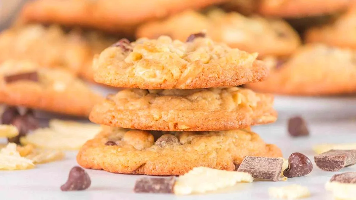 Three chocolate chip cookies are stacked on a white surface, with more cookies, chocolate chips, and cookie pieces scattered in the background.