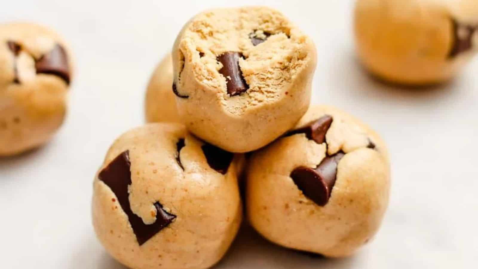 Four chocolate chip cookie dough balls are stacked on a white surface, with visible chocolate chunks embedded in the dough.