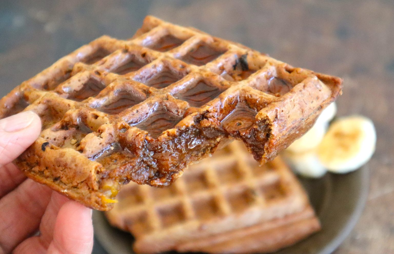 A hand holds a partially eaten waffle with melted chocolate, with more waffles and banana slices on a plate in the background.
