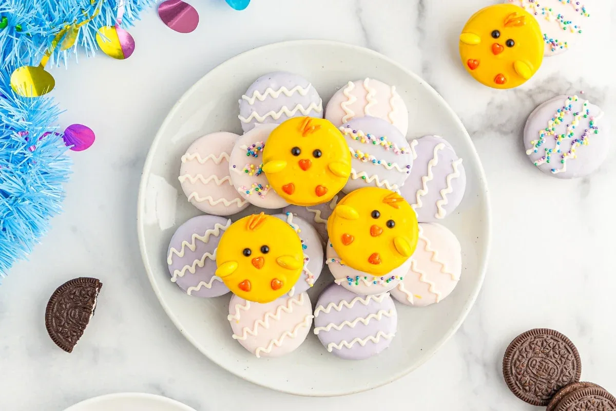 A plate of decorated cookies, including yellow chick faces and pastel-colored cookies with zigzag icing, sits on a white surface alongside Oreo cookies and colorful decorations.