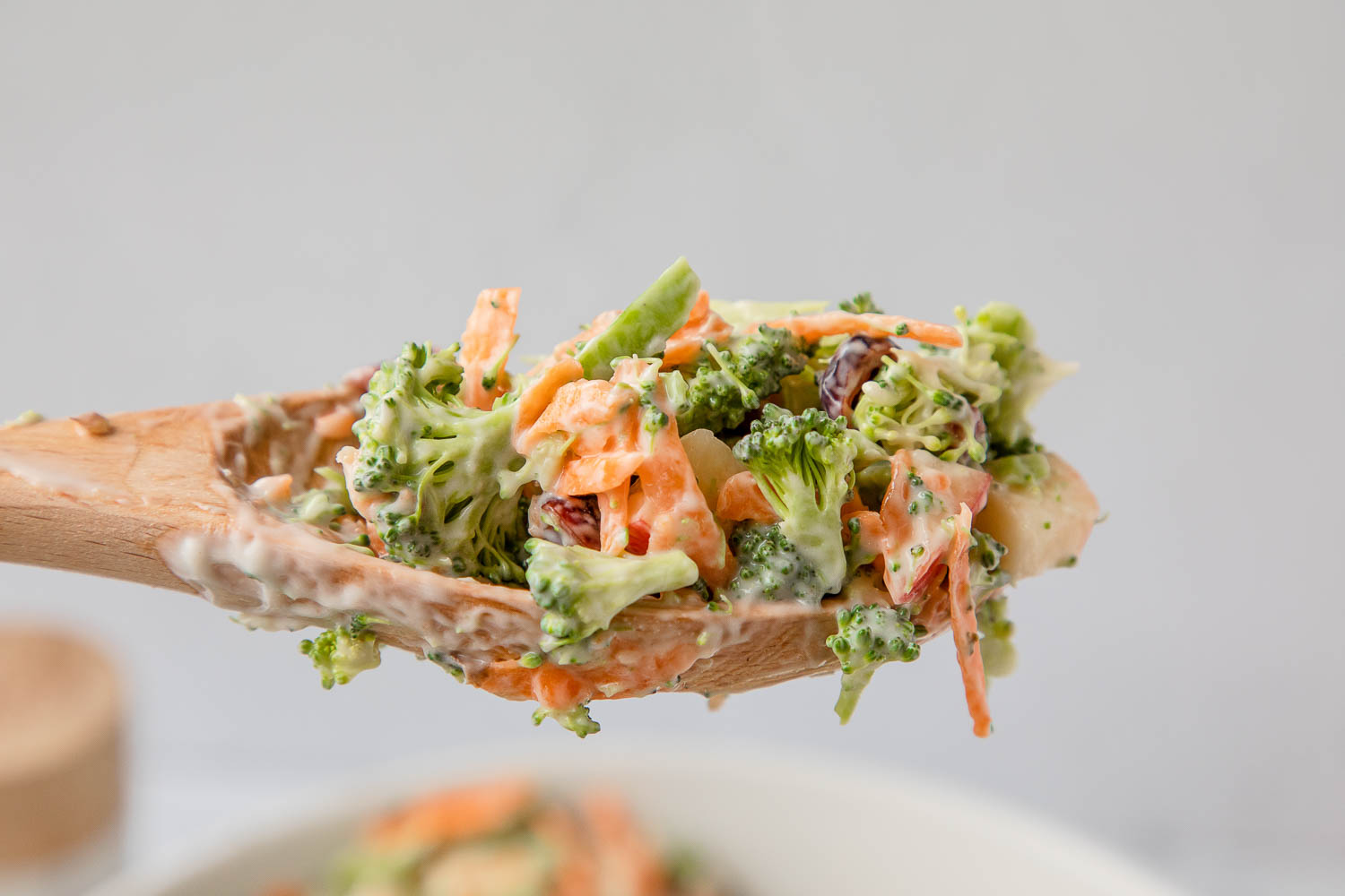 A wooden spoon holds a portion of broccoli salad with carrots, raisins, and a creamy dressing.