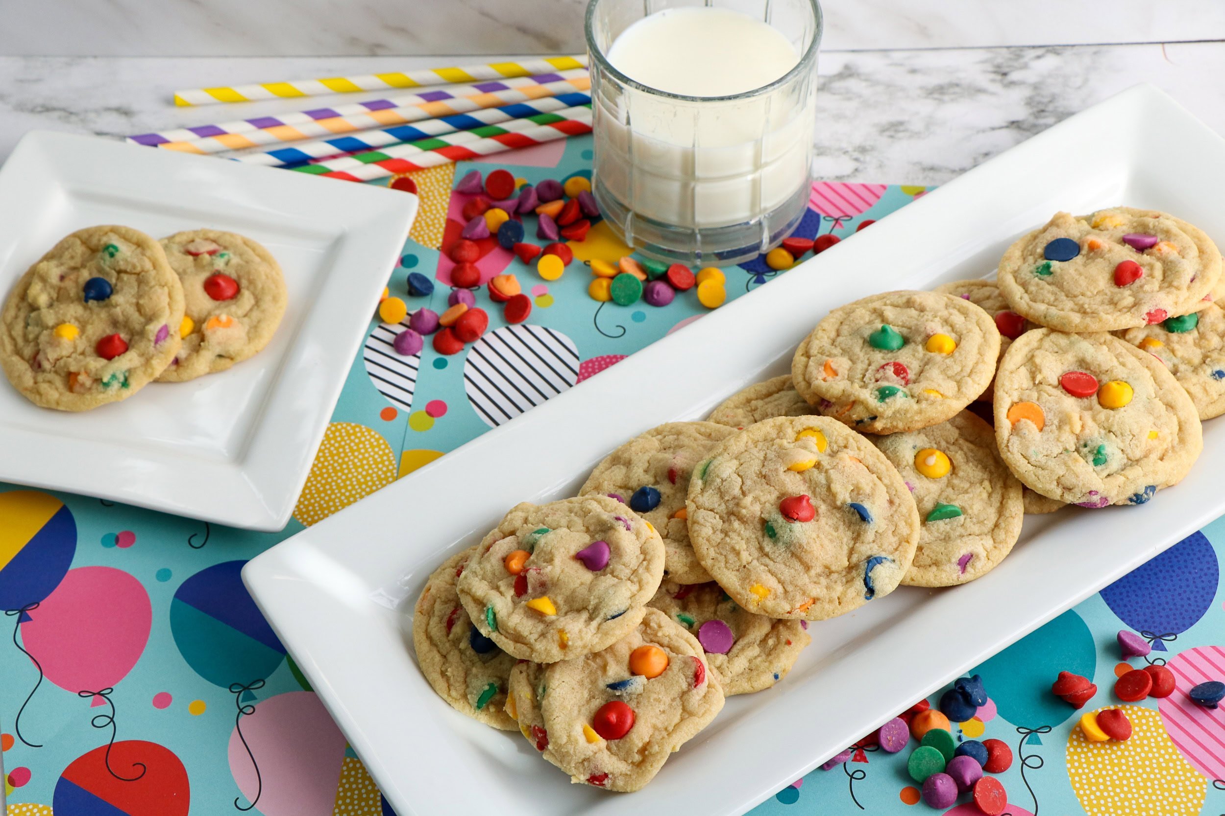 A plate of colorful candy-studded cookies sits next to a glass of milk and a smaller plate with more cookies, all on a festive balloon-themed table mat.
