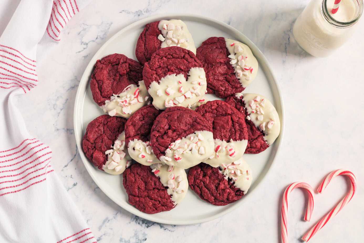A plate of red velvet cookies, some dipped in white chocolate and sprinkled with crushed peppermint, sits on a marble surface next to a bottle of milk and two candy canes.