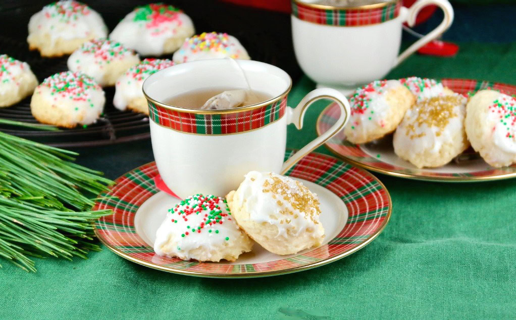 Teacup and saucer with plaid design, filled with tea, beside holiday cookies topped with white icing and colorful sprinkles, on a green tablecloth.