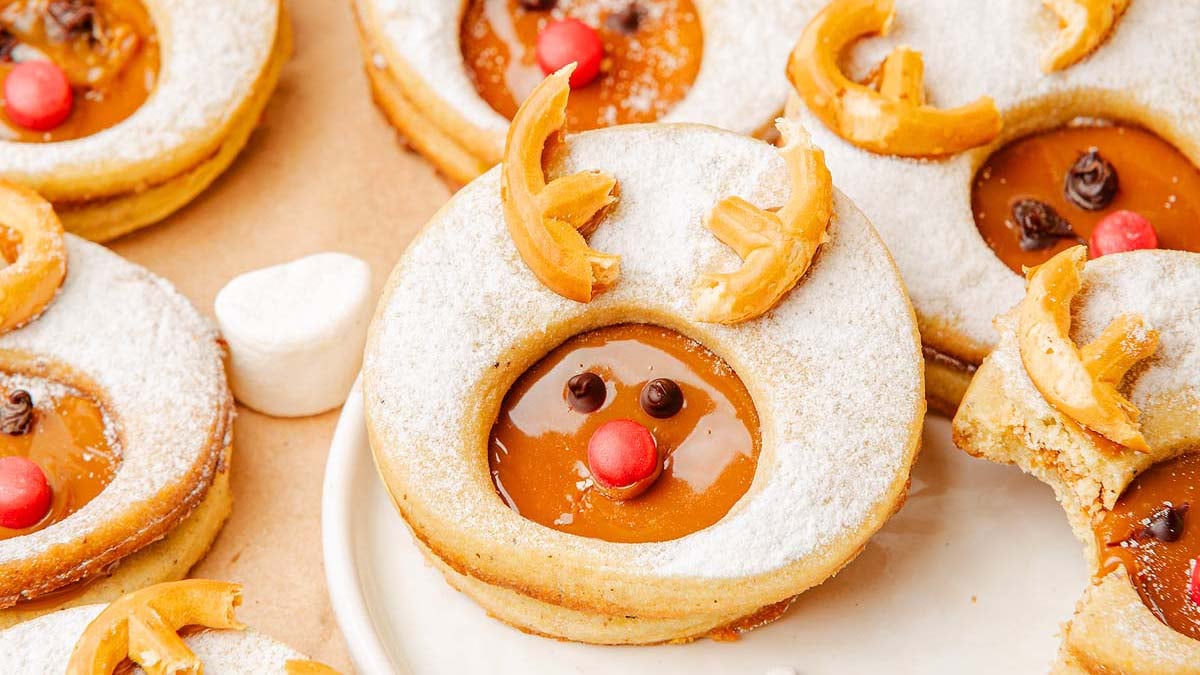 Round cookies with powdered sugar, caramel filling, and decorations resembling reindeer faces with pretzel antlers, candy noses, and chocolate eyes on a plate.