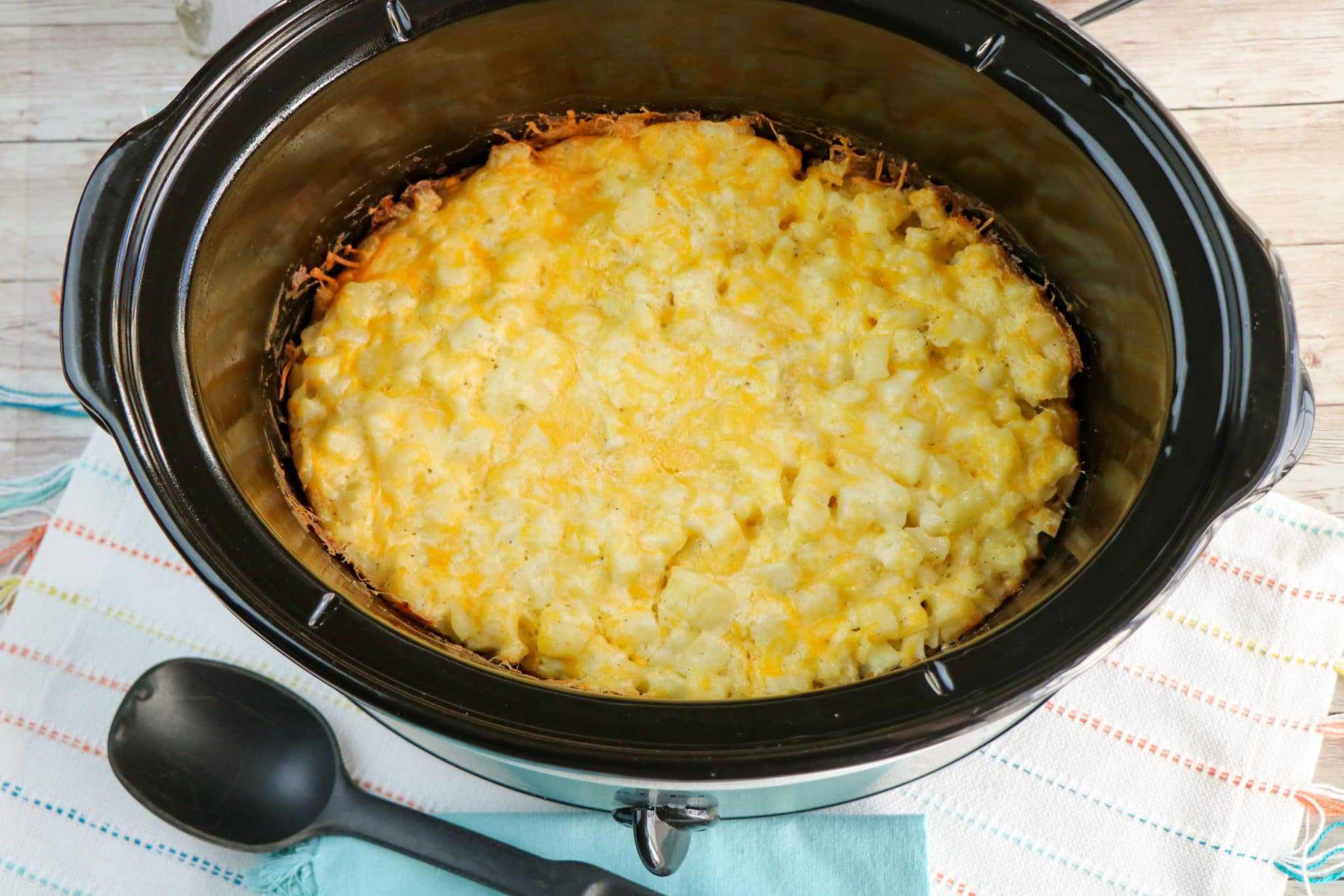 A slow cooker filled with a baked cheesy potato casserole, resting on a striped towel with a black serving spoon nearby.