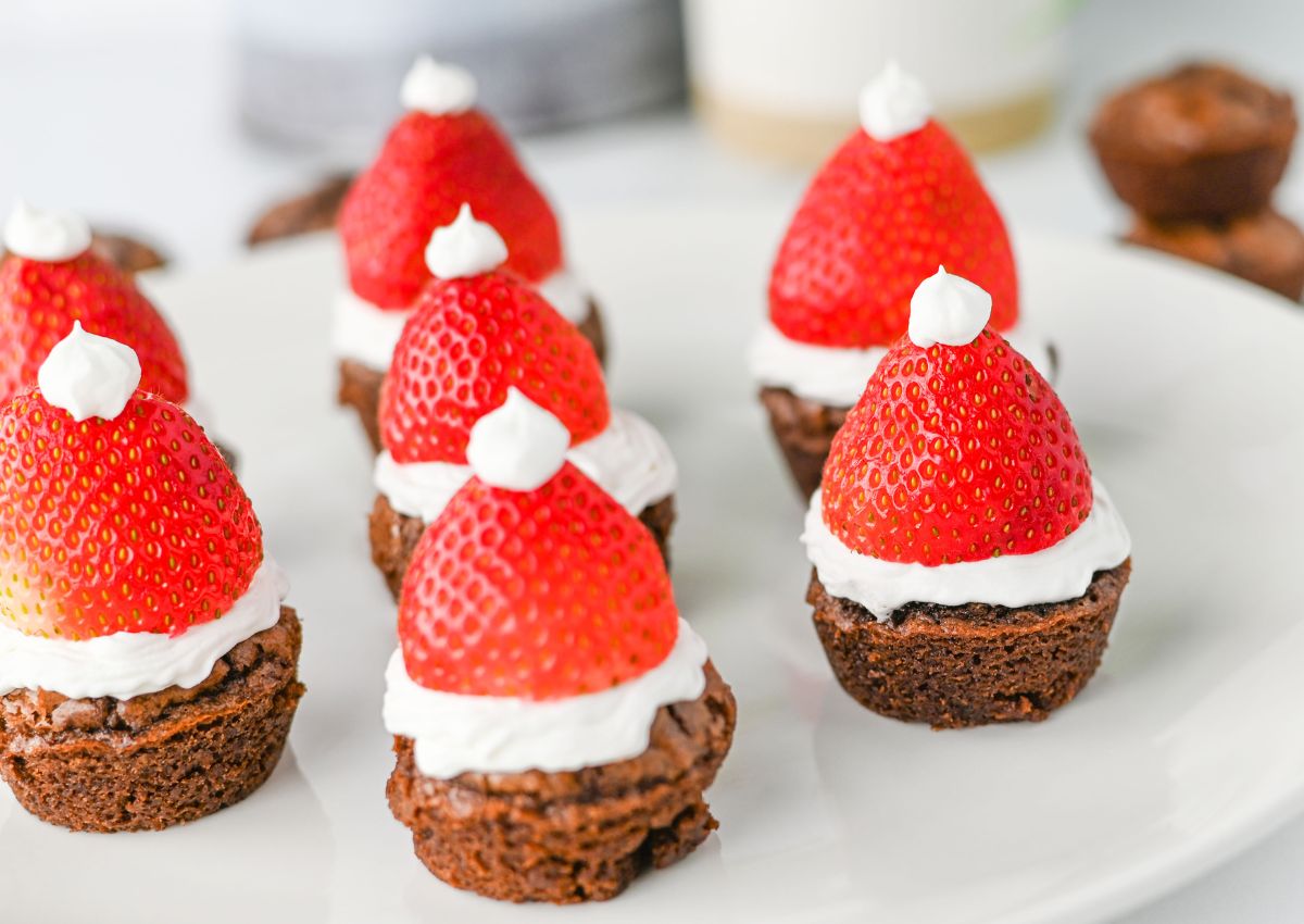 Chocolate brownie bites topped with whipped cream and strawberries arranged to look like Santa hats on a white plate.