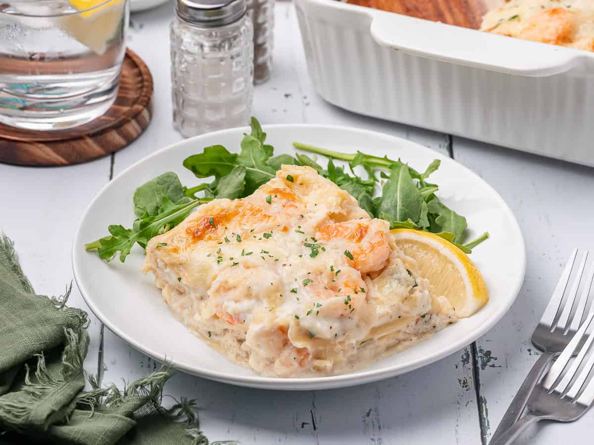 A serving of creamy seafood lasagna garnished with herbs, served with arugula and a lemon wedge on a white plate.