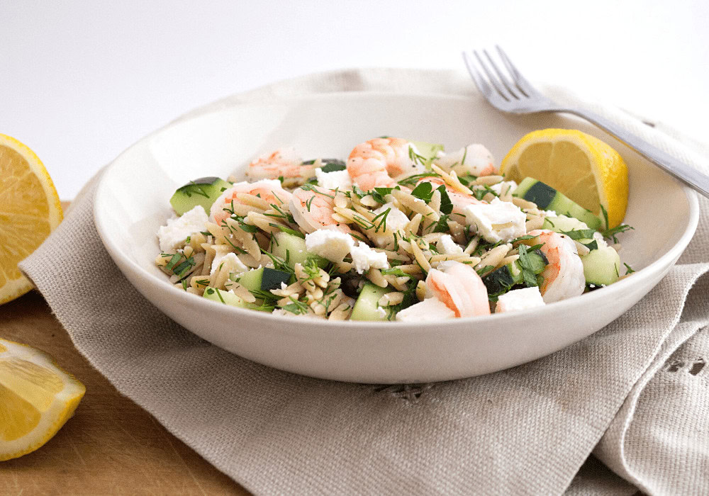 A bowl of pasta salad with shrimp, feta cheese, cucumber, and fresh herbs, garnished with lemon wedges, set on a neutral napkin with a fork beside it.