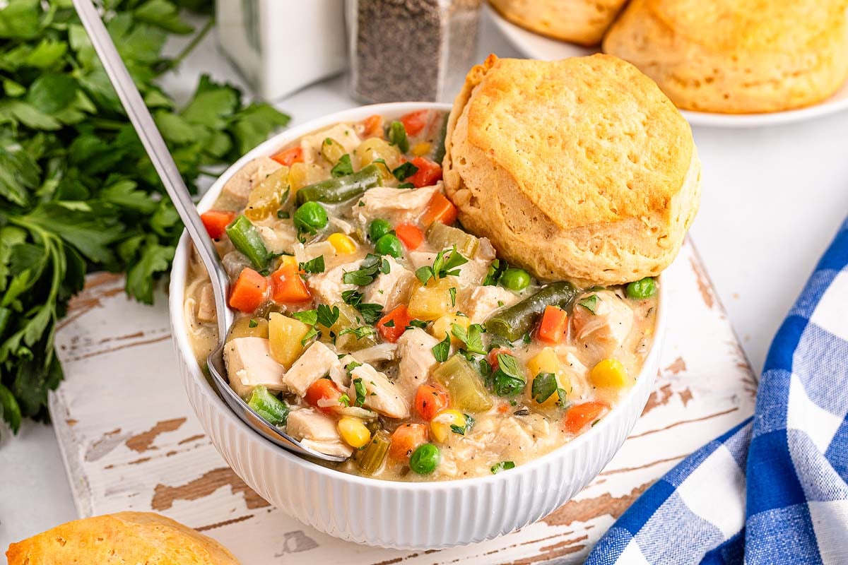 A bowl of creamy chicken and vegetable stew topped with a biscuit, with a spoon inside. Fresh parsley and biscuits are visible in the background.