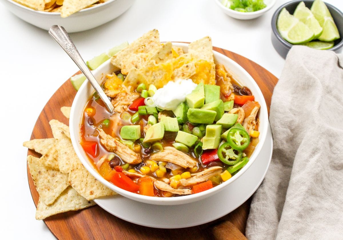 A bowl of tortilla soup with shredded chicken, corn, beans, diced avocado, jalapenos, and sour cream, garnished with tortilla chips. Lime wedges and green onions on the side.
