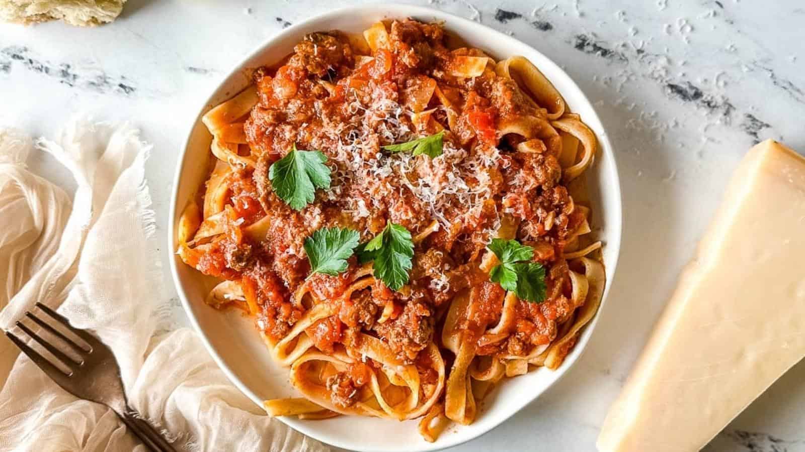 A bowl of tagliatelle pasta topped with meat sauce, grated cheese, and parsley, placed on a marble surface next to a block of cheese and a fork.