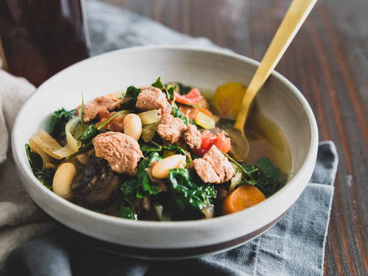 A bowl of soup containing chunks of meat, white beans, leafy greens, carrots, and other vegetables, with a spoon inside the bowl.