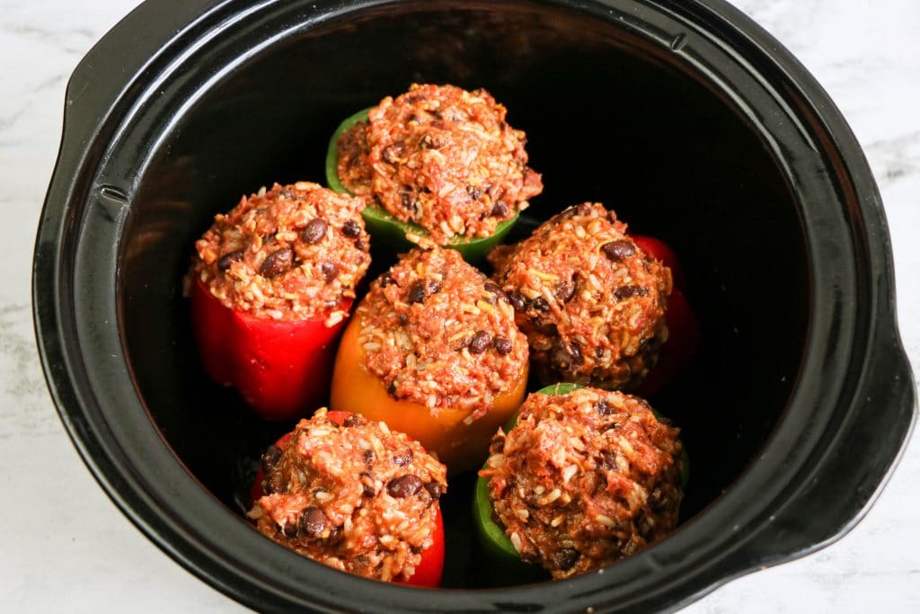 Six bell peppers stuffed with a rice and bean mixture are arranged upright inside a black slow cooker before being cooked.