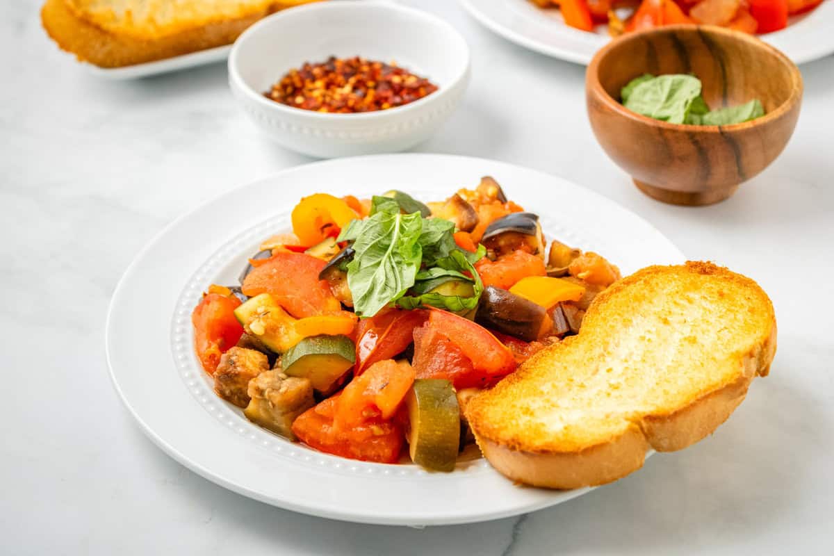 A plate of ratatouille with sliced zucchini, tomatoes, and eggplant, garnished with basil, served with a slice of toasted bread. Bowls of chili flakes and basil are in the background.