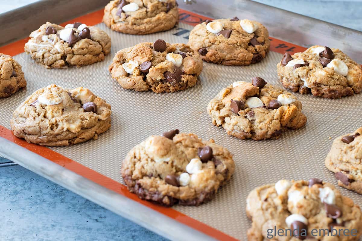 A baking tray lined with a silicone mat holds freshly baked cookies with chocolate chips and marshmallows.