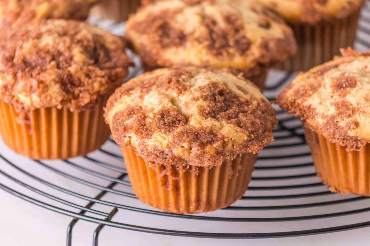 Four cinnamon streusel muffins cool on a black wire rack, showing a crumbly topping and golden-brown texture.