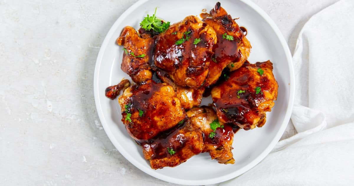 Five glazed, cooked chicken thighs garnished with parsley are arranged on a white plate, set on a light-colored surface.