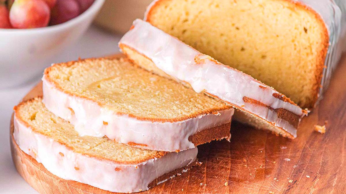 A loaf of glazed pound cake is sliced on a wooden cutting board, with two slices lying flat in front of the remaining loaf.