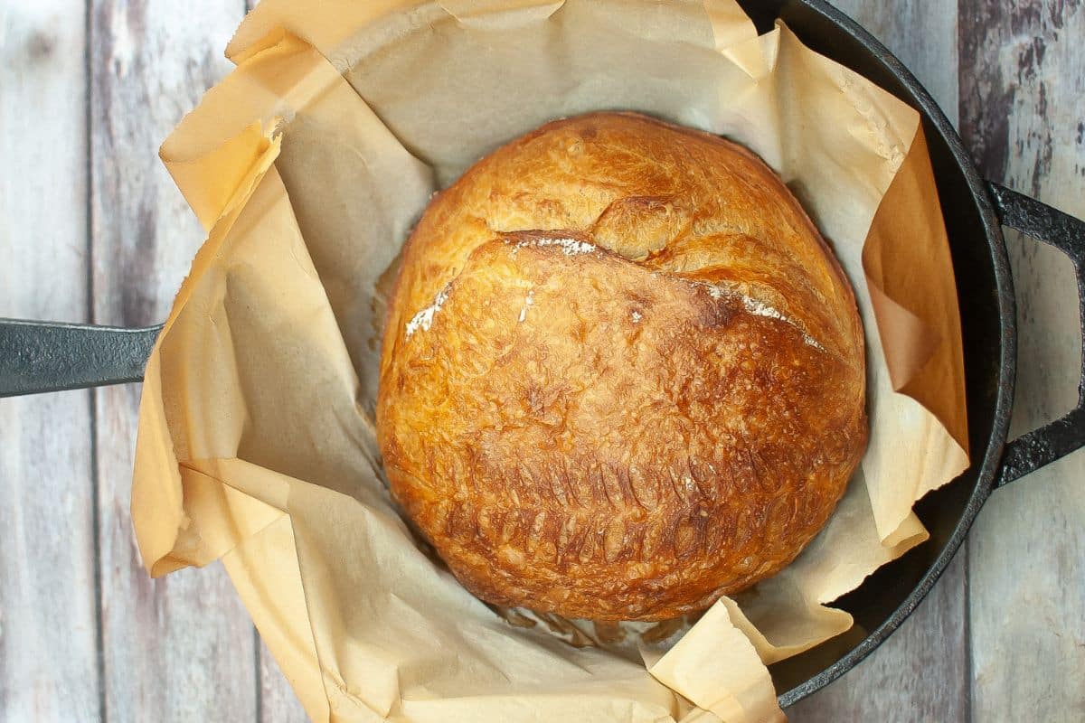 A round loaf of golden-brown bread on parchment paper inside a black cast iron pot, viewed from above.
